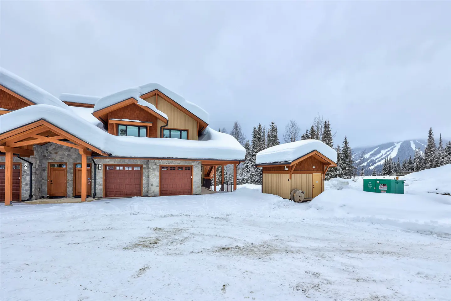 A snow-covered house with brown garage doors and a small shed in a snowy landscape.