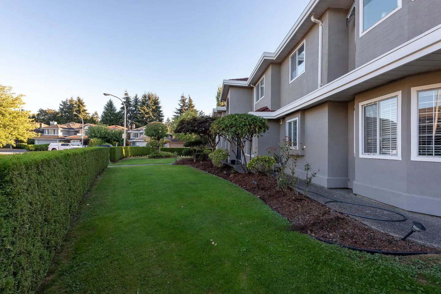Side view of a grey two-story townhouse with a manicured lawn, tall hedge, and landscaped garden beds under a blue sky.