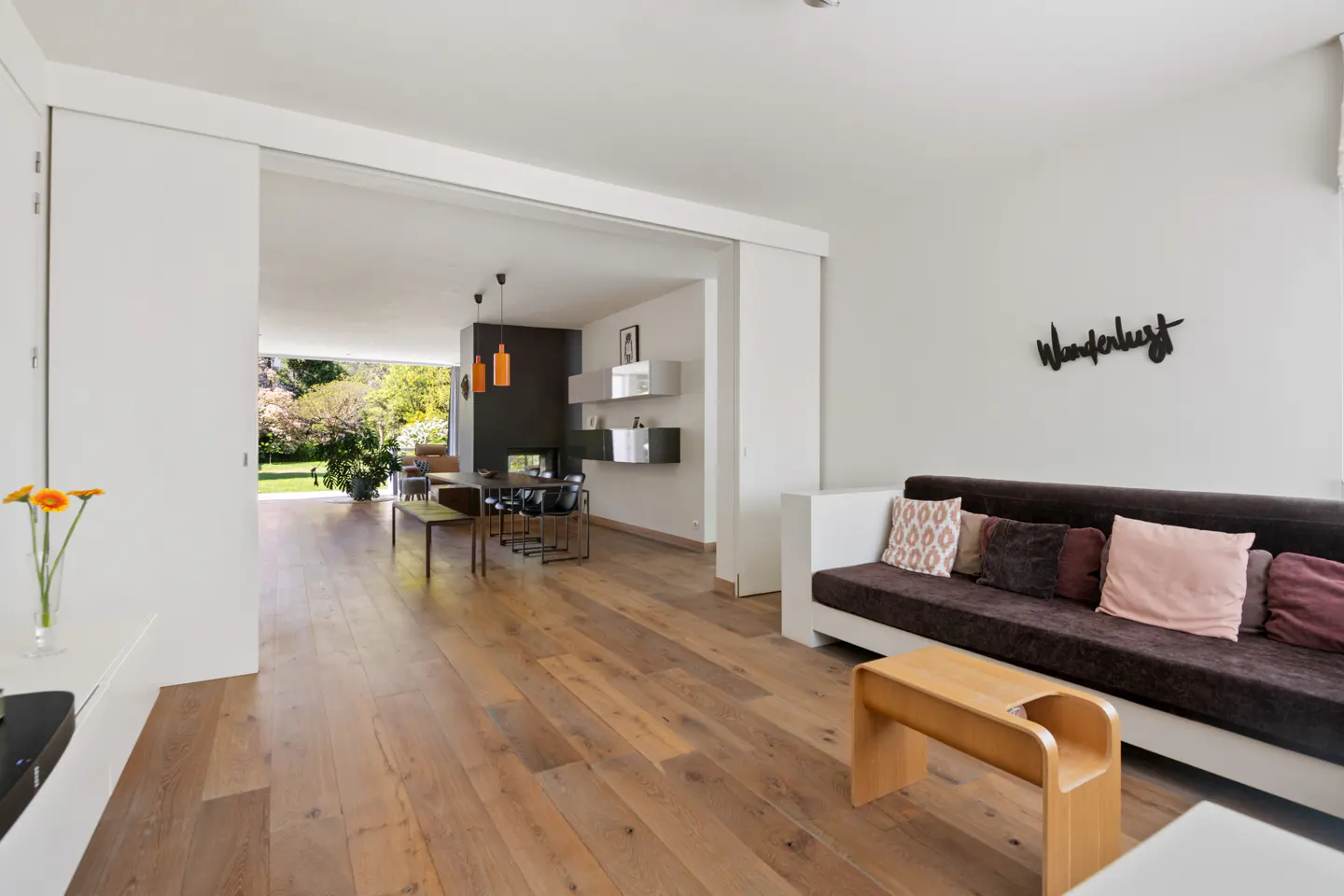 A bright, modern living room with hardwood floors, a brown sofa with pillows, and an open doorway to a dining area and garden.