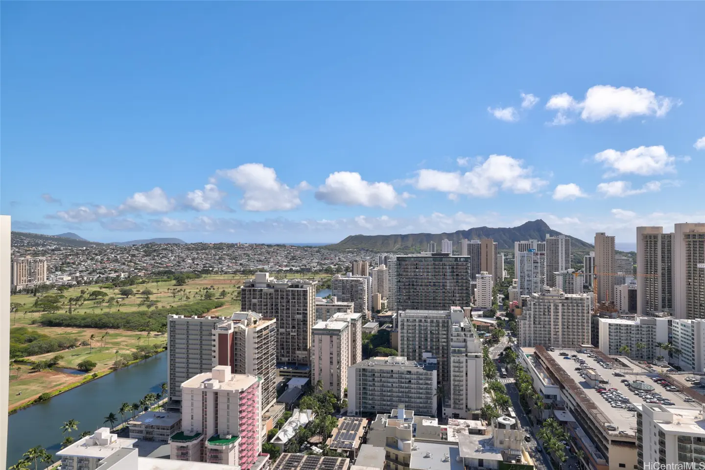 Cityscape view of Honolulu, Hawaii, with Diamond Head in the background under a blue sky with scattered clouds. Tall buildings and a canal are visible.