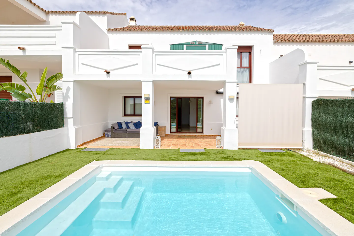 White villa with a private pool, artificial grass lawn, and a covered patio featuring a sofa and open glass doors.