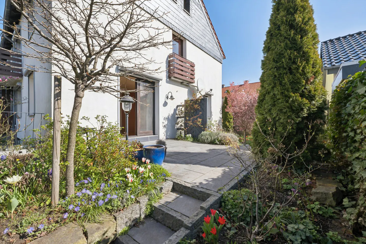 Exterior view of a white house with a gray roof, stone patio, and garden with flowers and trees.