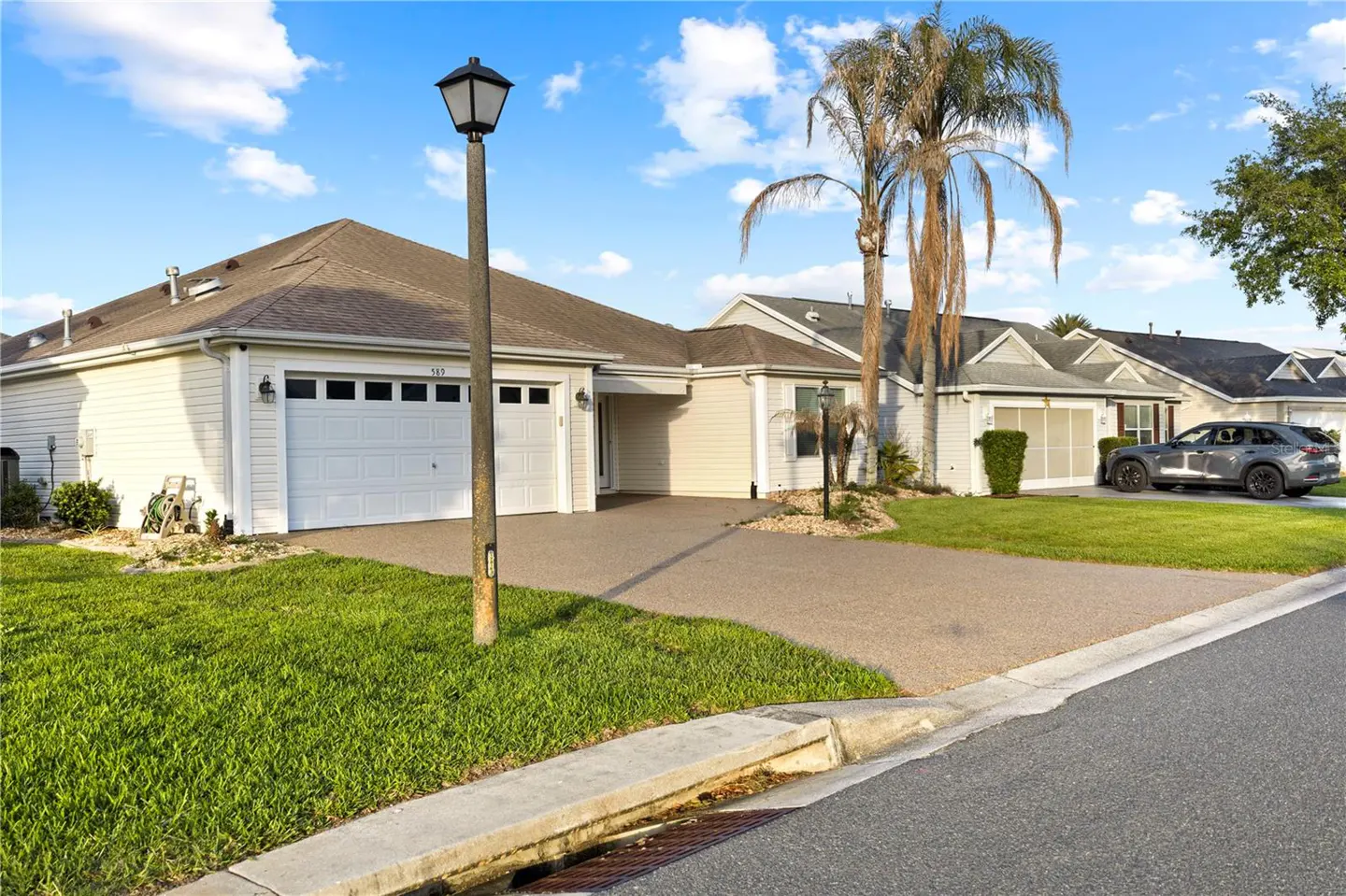 Exterior view of a one-story home with a white garage door, brown roof, green lawn, and palm trees under a blue sky.