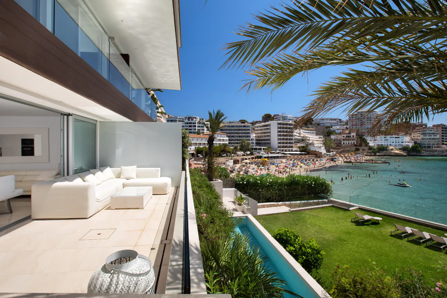 Balcony view of a beach with white sofas, green lawn, and blue water. Palm trees frame the sunny scene.