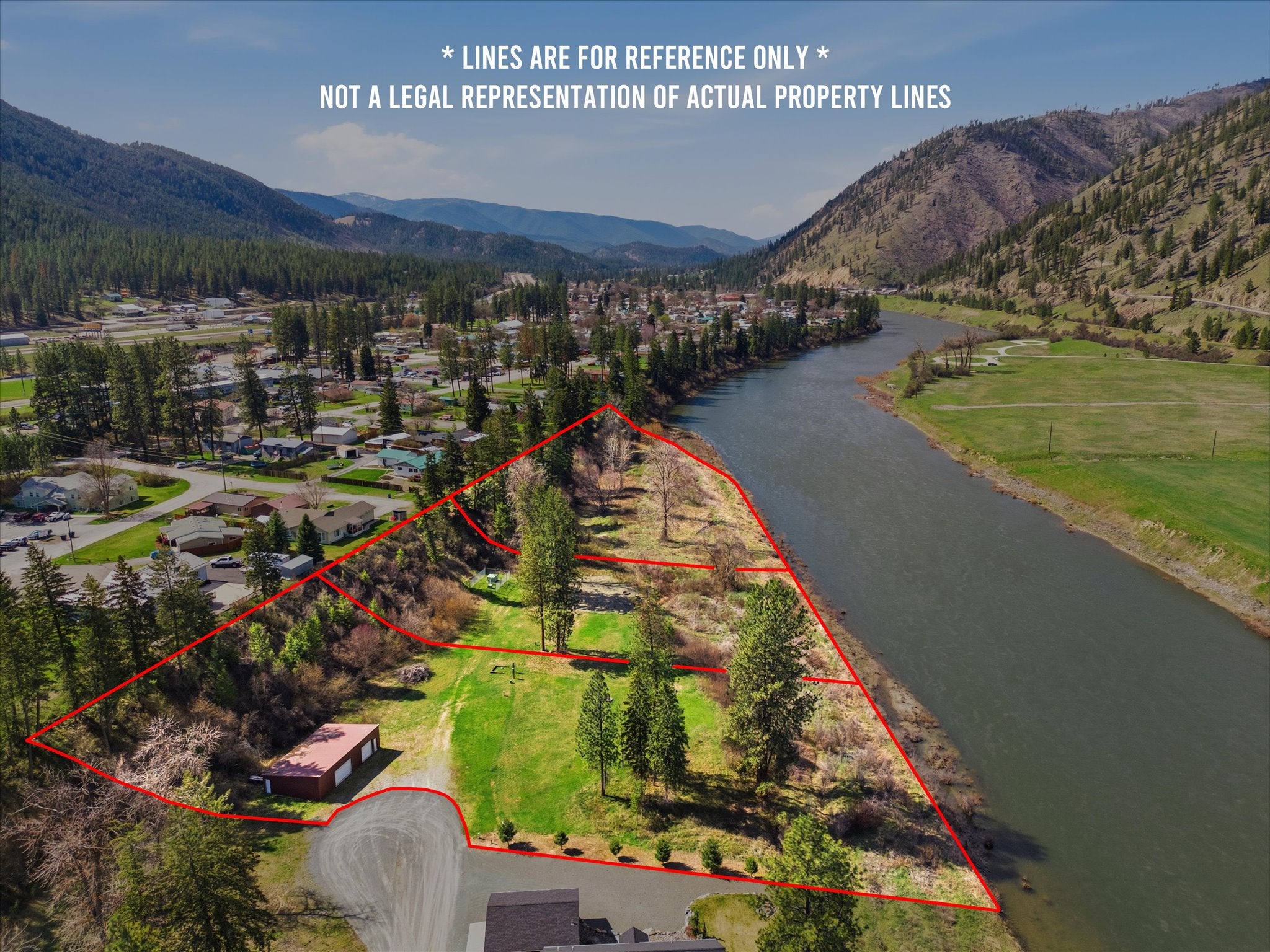 Aerial view of a riverfront property lot outlined in red, featuring trees, a small building, and nearby town and mountains. [AI-generated text]