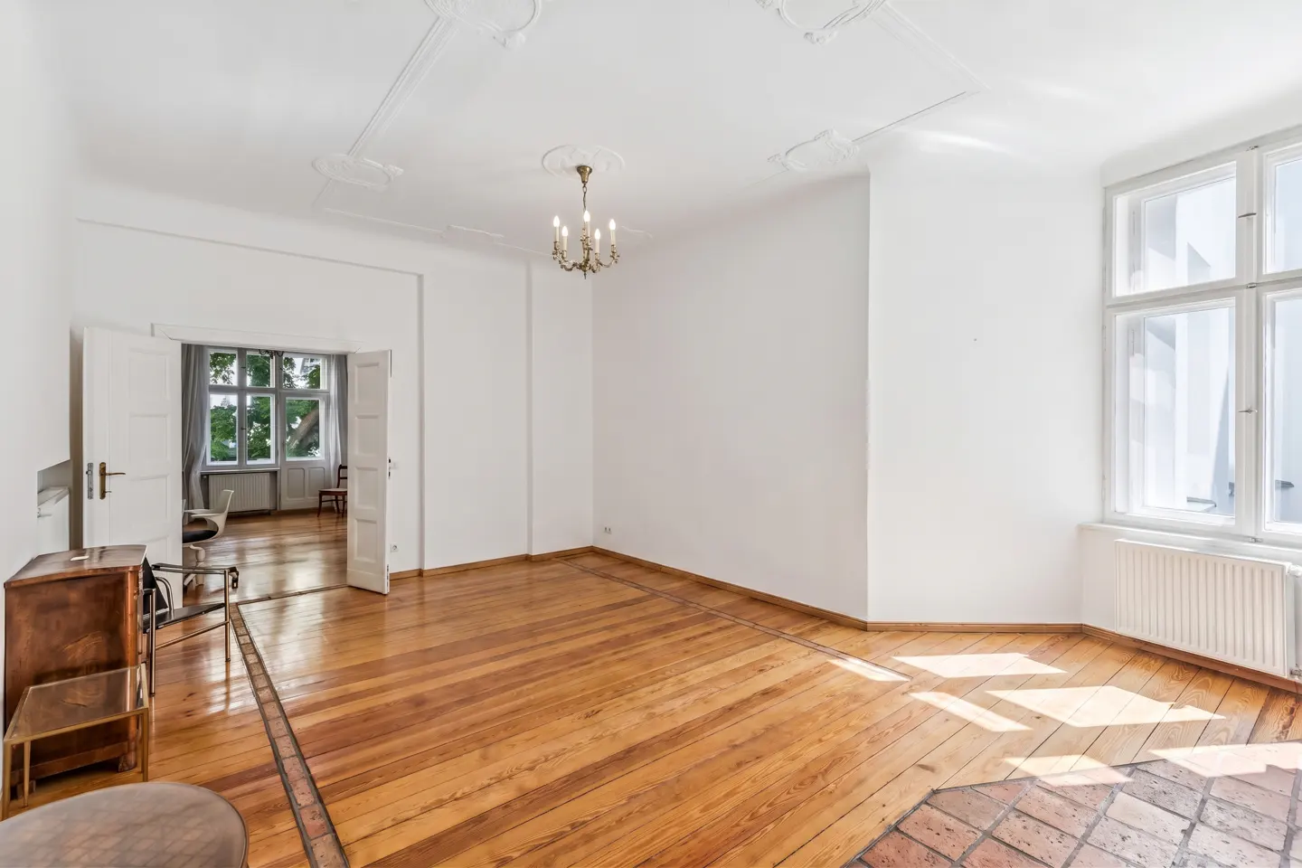 Bright, empty room with white walls, ornate ceiling molding, a small chandelier, and polished hardwood floors.