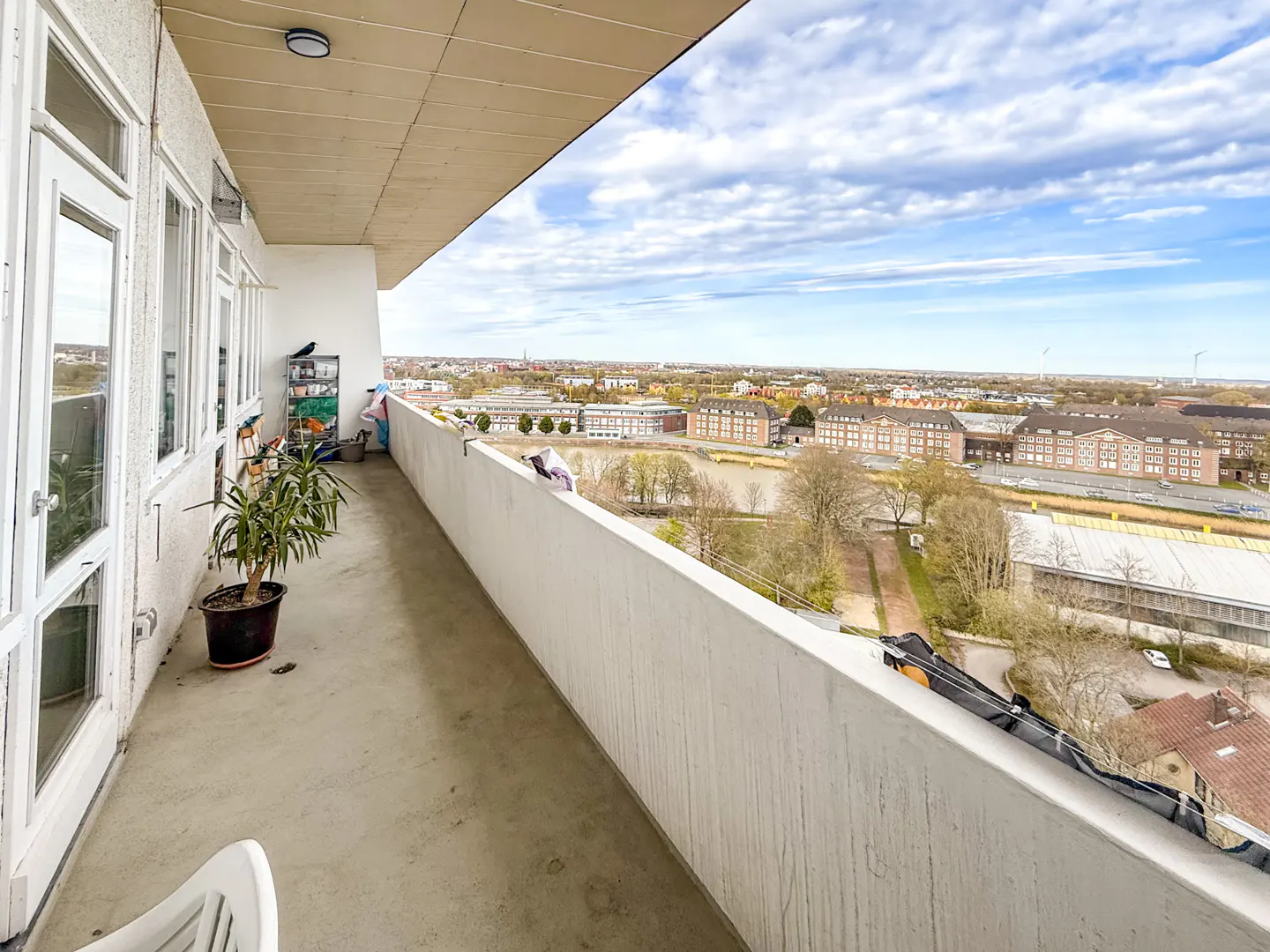 A high-angle view from a balcony shows a city skyline under a cloudy sky. A potted plant and storage rack sit near white-framed windows.