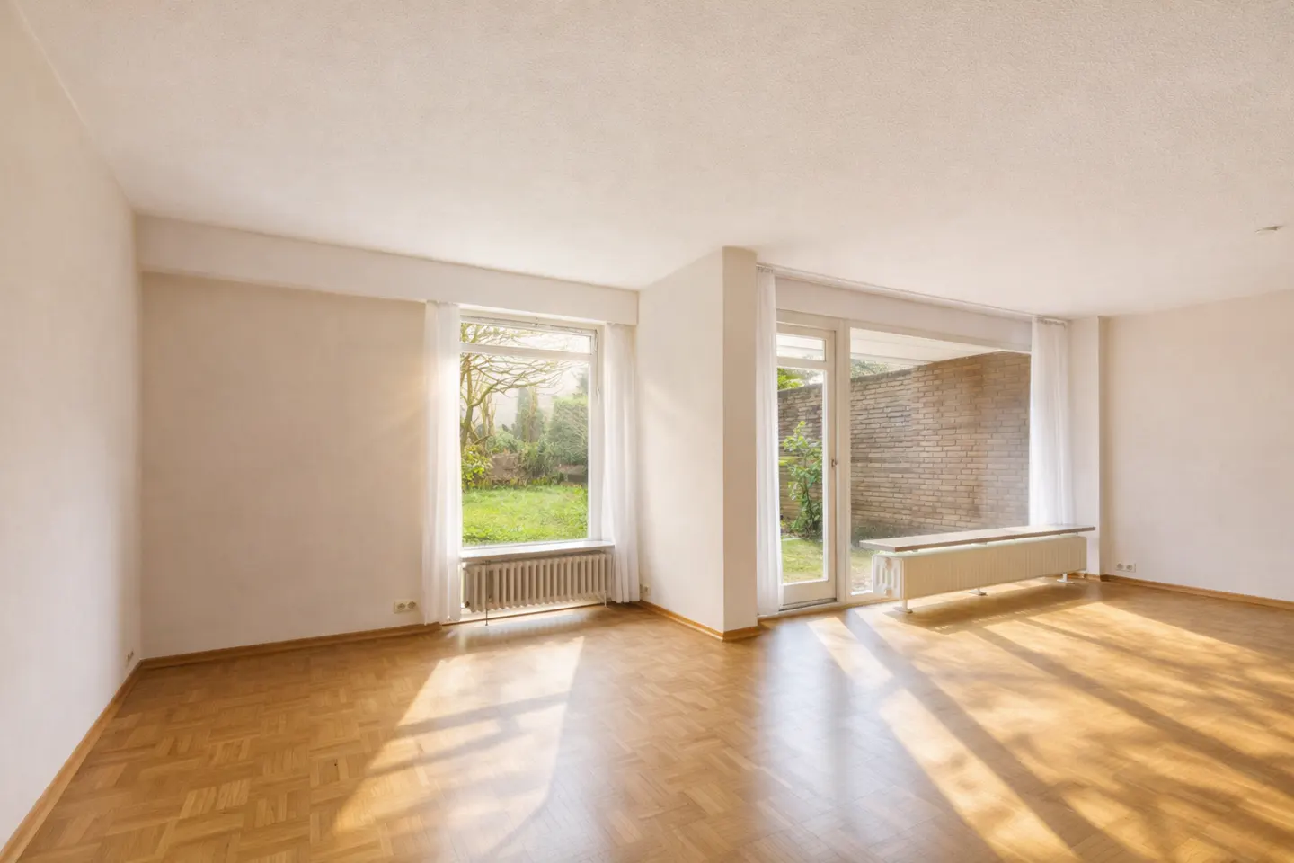 Bright, empty room with parquet floors, white walls, and large windows overlooking a green garden and brick patio.