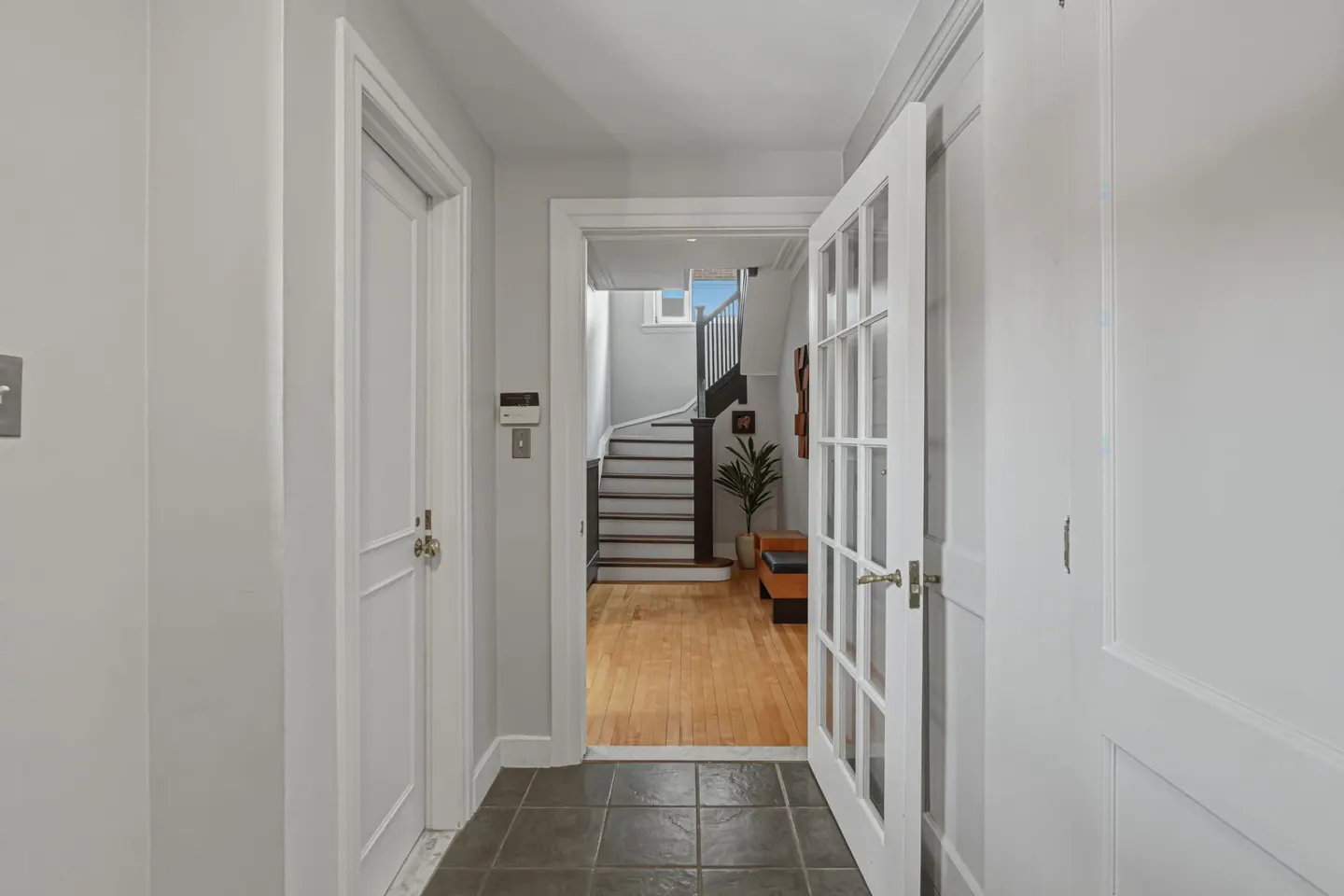 Hallway with gray tile floor, white walls, and open white French doors leading to a room with hardwood floors and stairs.
