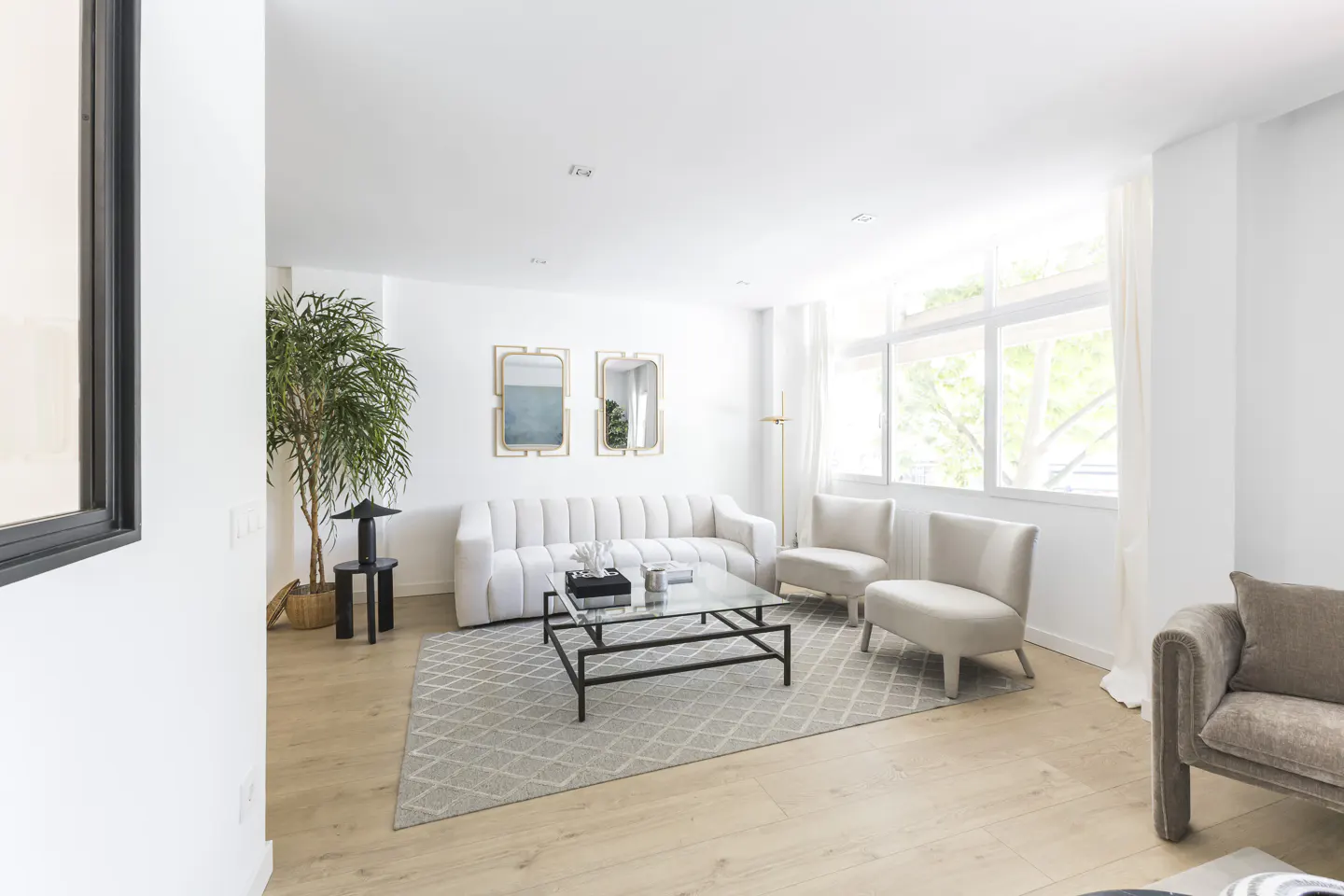 Bright living room with white walls, light wood floors, and large windows. A white sofa, two chairs, and a glass coffee table sit on a gray patterned rug.