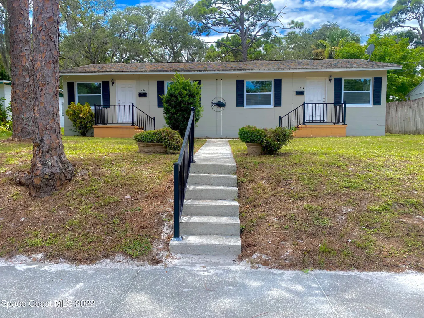 A tan duplex with black shutters and railings, a concrete walkway, and green lawn on a sunny day.