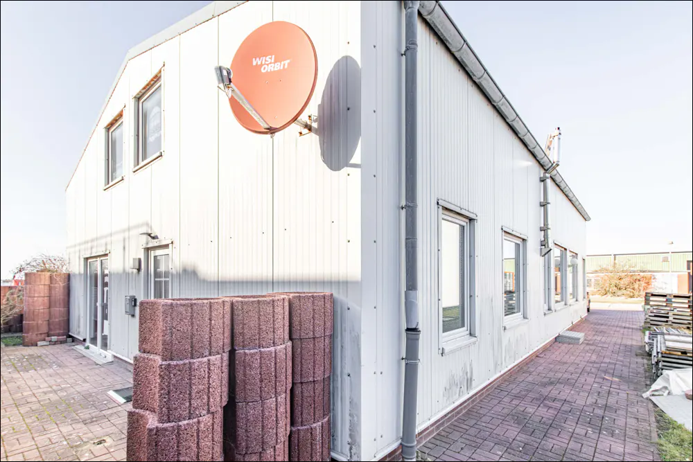 Exterior view of a two-story white building with a red satellite dish. Piles of red bricks sit on a brick-paved walkway.