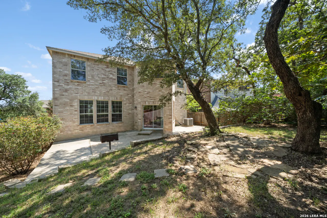 Backyard view of a two-story tan brick house with a patio, grill, and large trees providing shade on a sunny day.