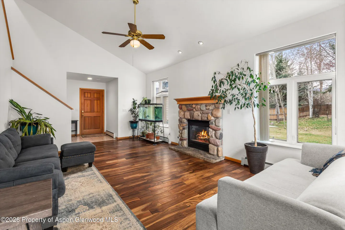 Living room with hardwood floors, white walls, and a stone fireplace with a wood mantel. A ceiling fan hangs above. A large window overlooks a green lawn.