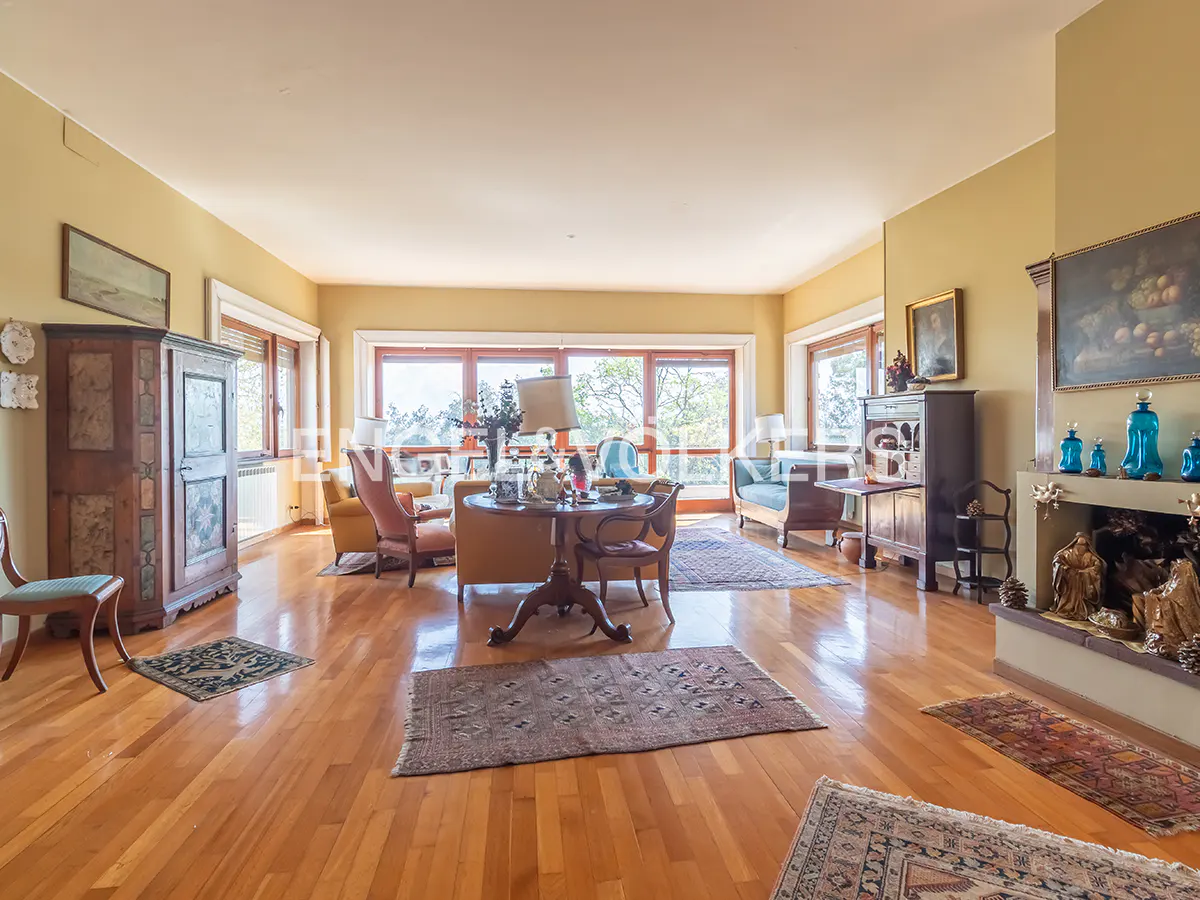 Spacious living room with hardwood floors, rugs, and large windows. Furniture includes sofas, chairs, and antique cabinets. Fireplace with blue vases.