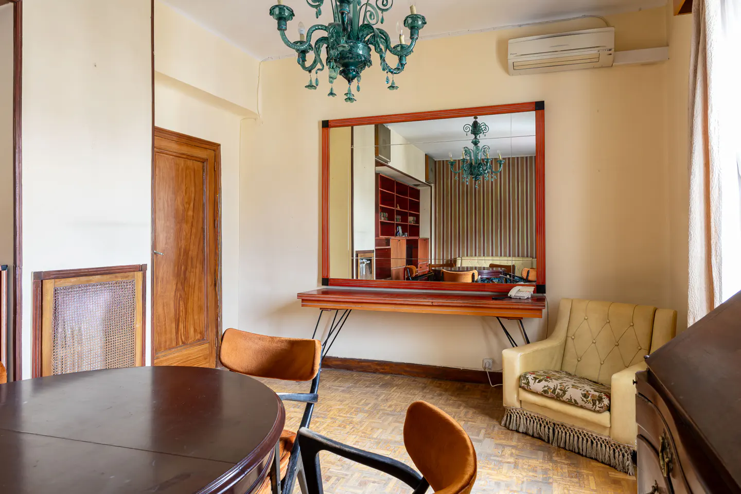 A vintage-style room with a dark wood table, orange chairs, and a cream armchair. A large mirror reflects a bookshelf and green chandelier.