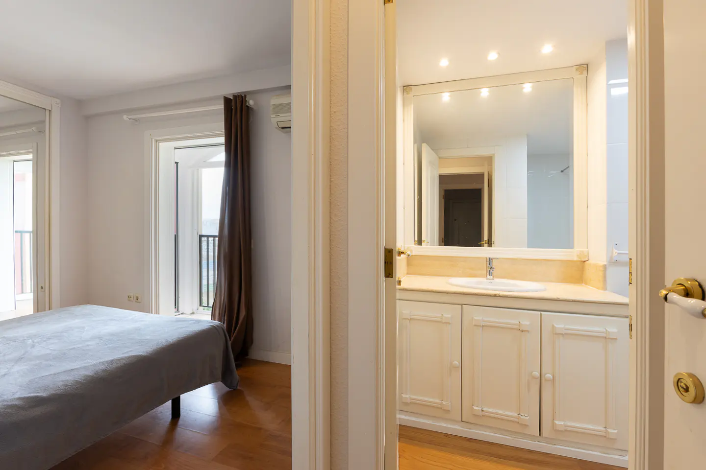 View into a bedroom with a gray bedspread and a bathroom with a white vanity and mirror.
