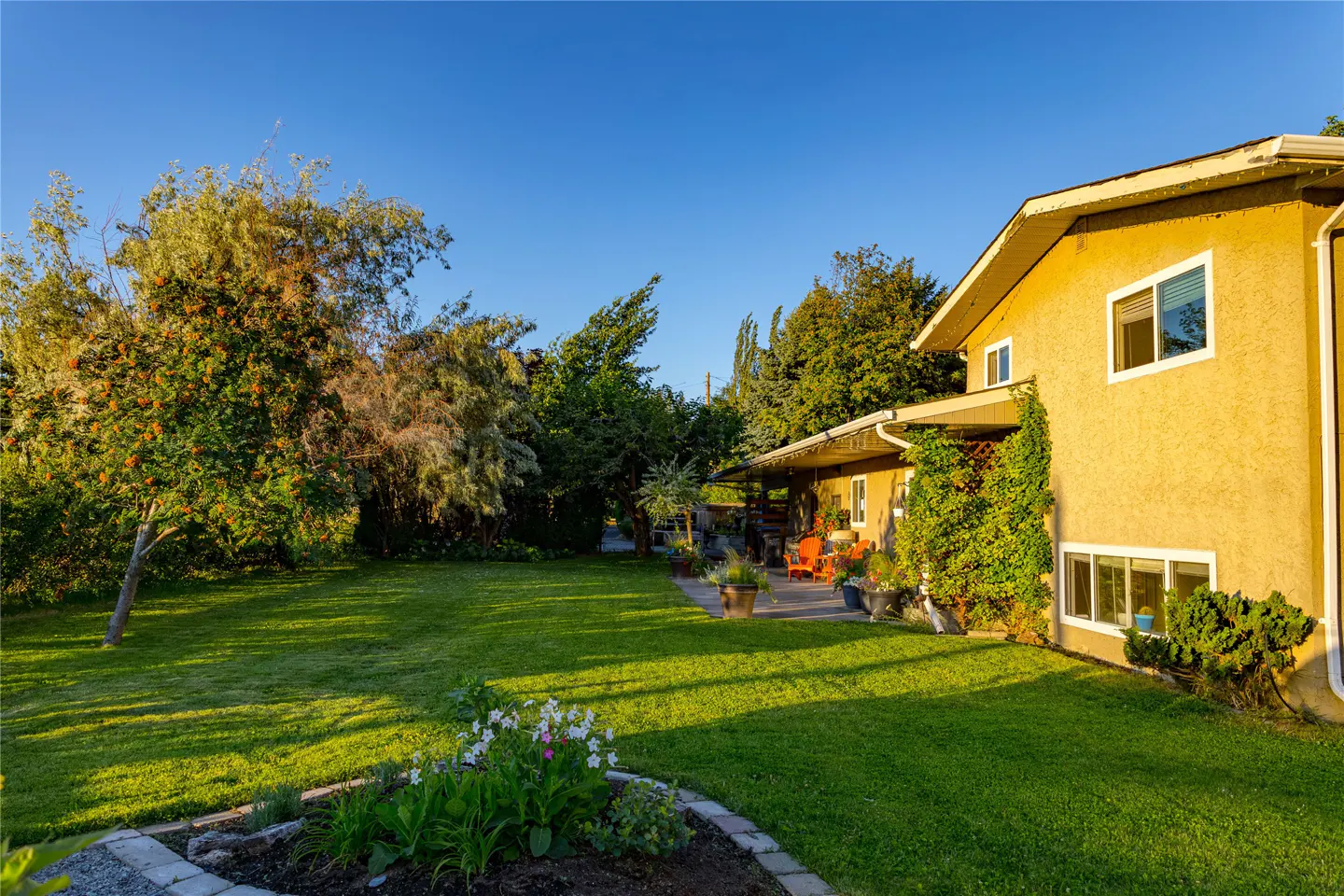 A yellow two-story house with a green lawn and trees under a blue sky. A patio with orange chairs is visible.