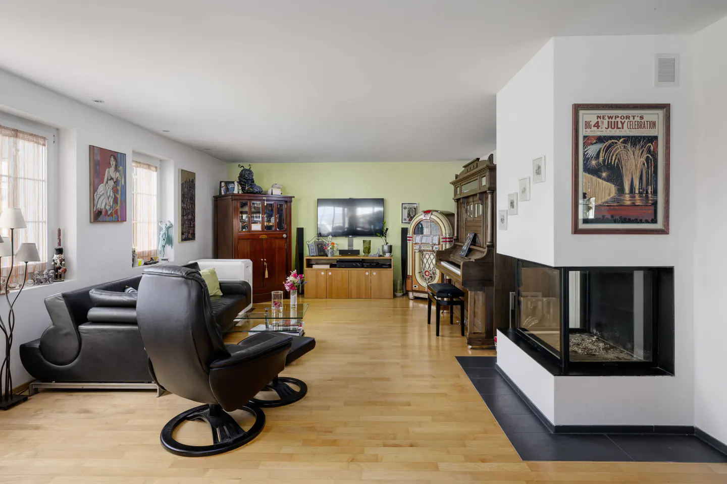 Bright living room with wood floors, black leather furniture, and a white fireplace. A jukebox and piano stand near a wall with a "July 4th" poster.