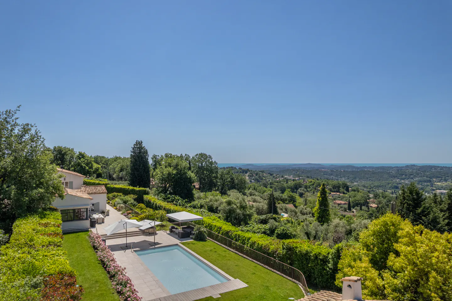 Aerial view of a luxury home with a pool, green lawn, and lush trees under a clear blue sky.