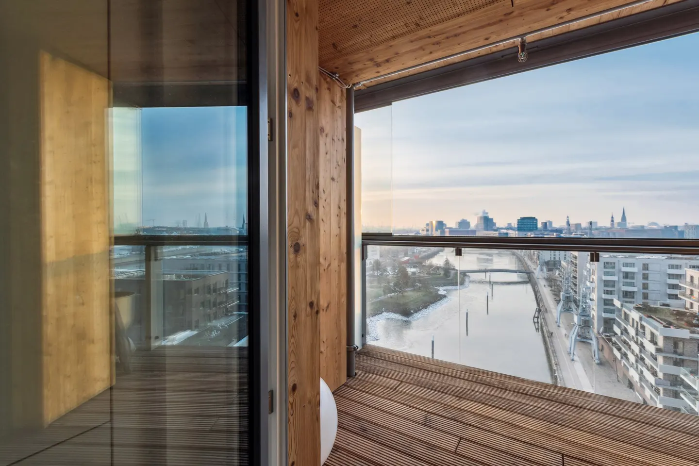 Balcony view: Wood deck, glass railing, city skyline. River below, modern buildings. Wood paneled walls. Sky is blue with some clouds.