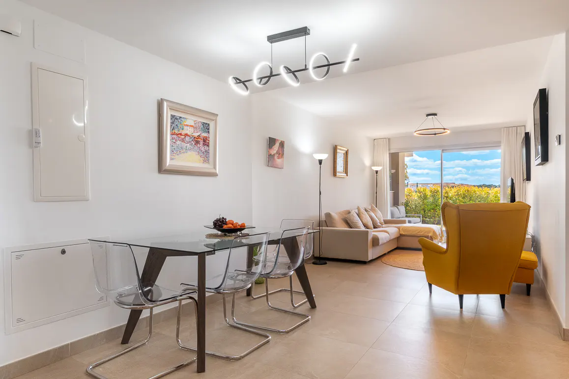 Bright, modern apartment interior with a glass dining table, yellow armchair, and a view of trees through a large window.