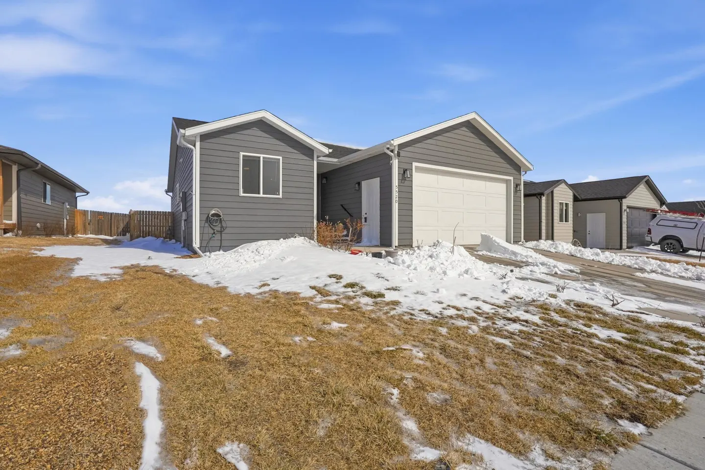 Single-story gray house with a white garage door and snow-covered yard under a bright blue sky.