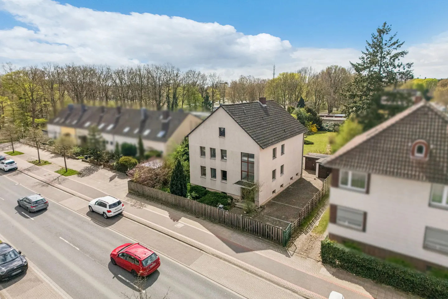 Aerial view of a cream two-story house with a gray roof, surrounded by a wooden fence and greenery, next to a street with cars.