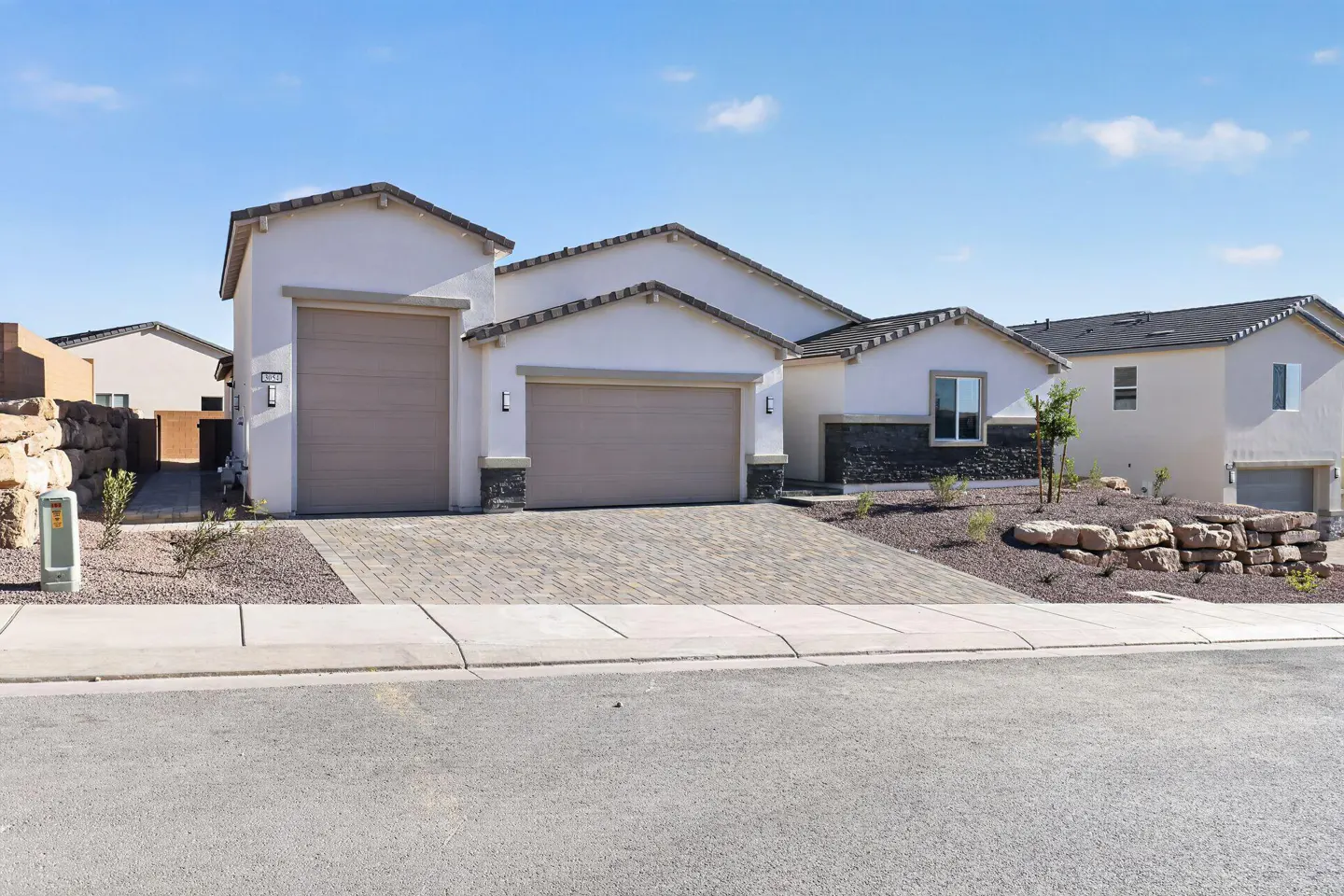 A modern, single-story home with a beige exterior, two-car garage, and a brick driveway under a clear blue sky.