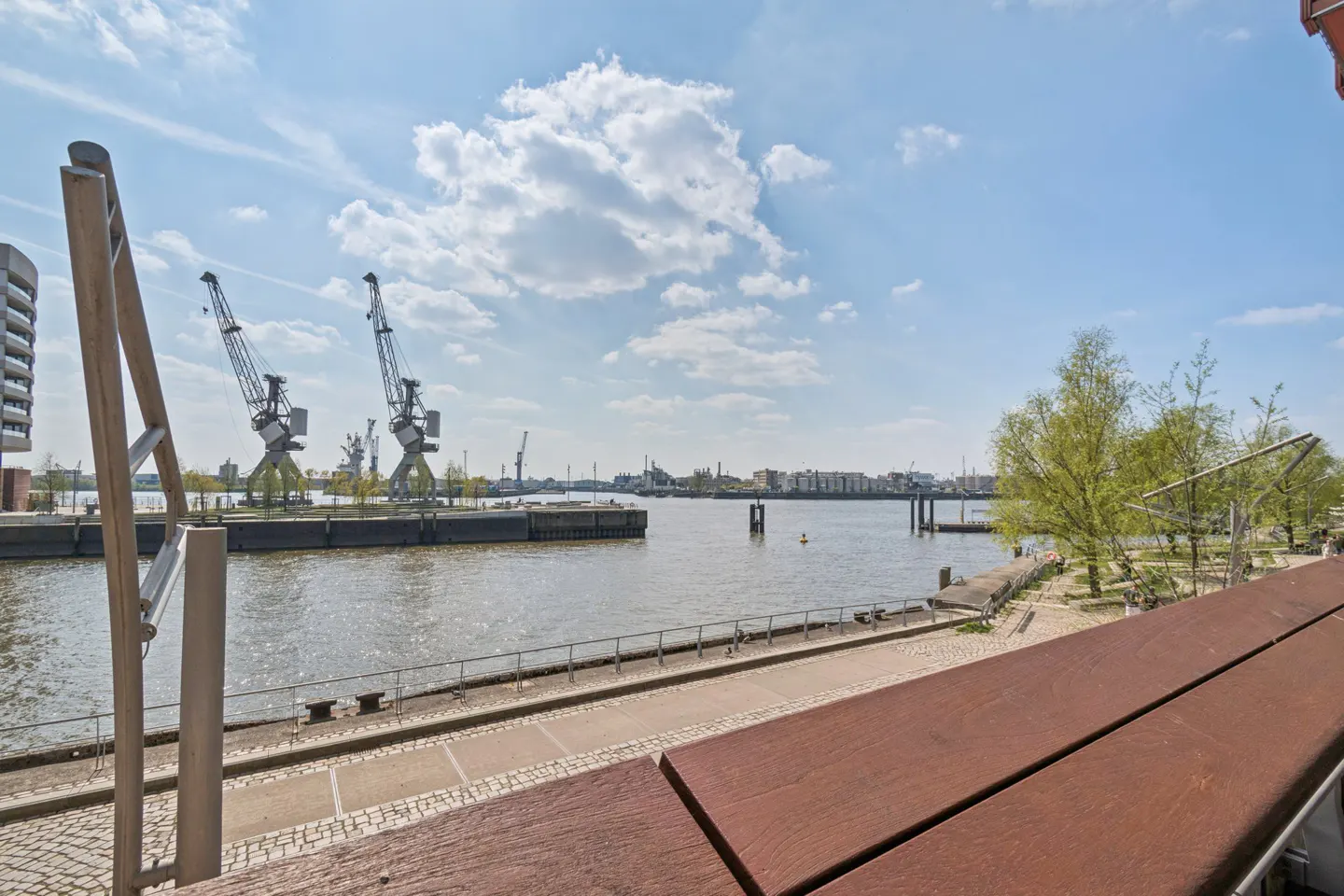 View of a harbor with cranes, water, and a blue sky with clouds. A wooden railing is in the foreground.