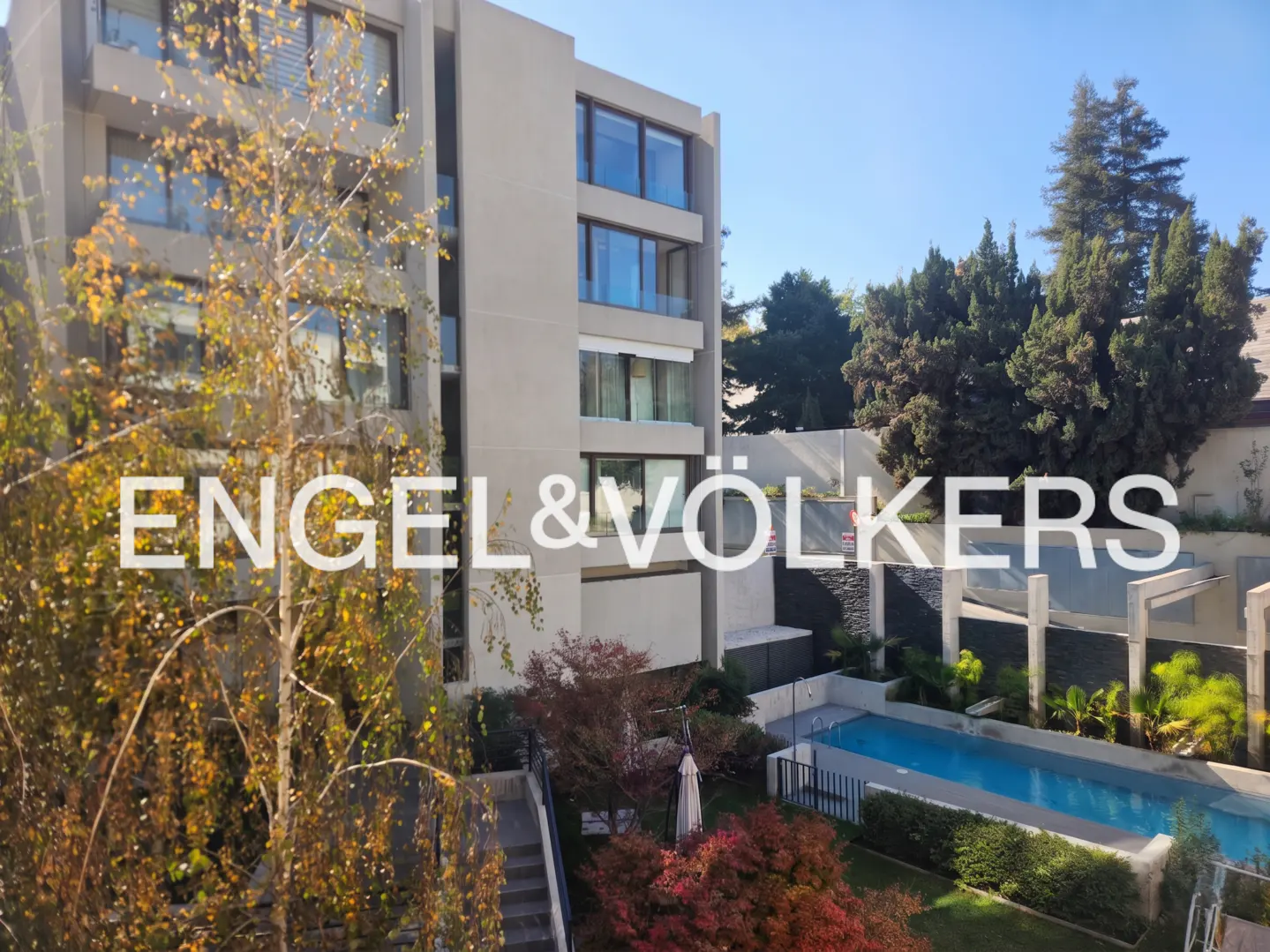 Exterior view of a modern apartment building with a pool and garden, under a blue sky. Engel & Völkers logo overlay.