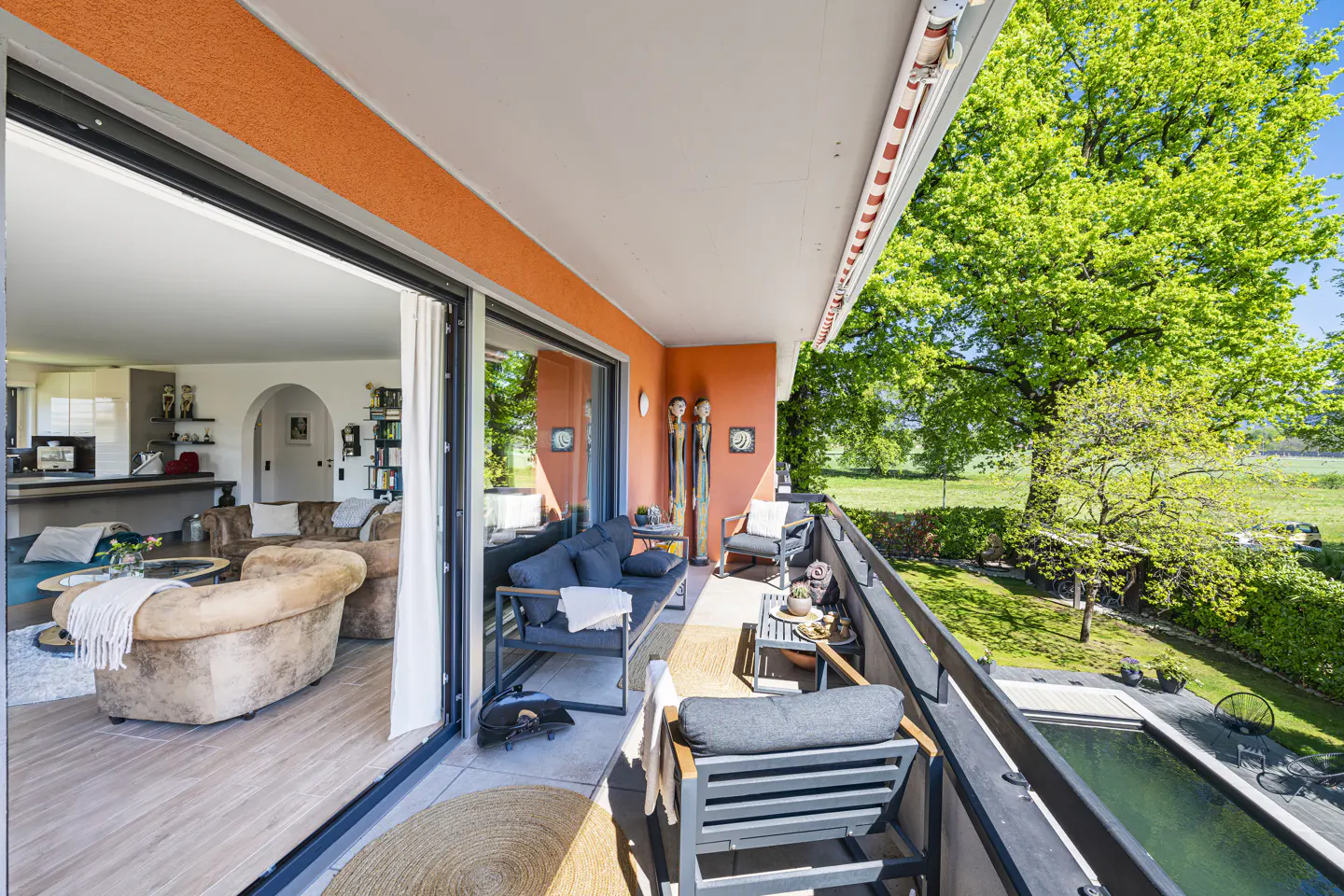 A bright, orange balcony with outdoor seating overlooks a green lawn and pool. Sliding glass doors open to a living room with a brown sofa.