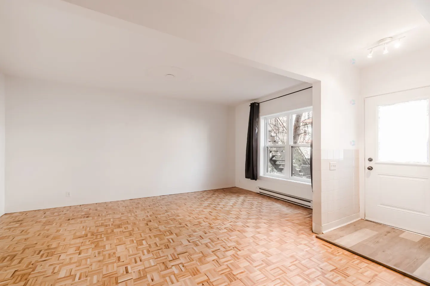 Bright, empty room with parquet floors, white walls, and a window with a black curtain. A white door is on the right.