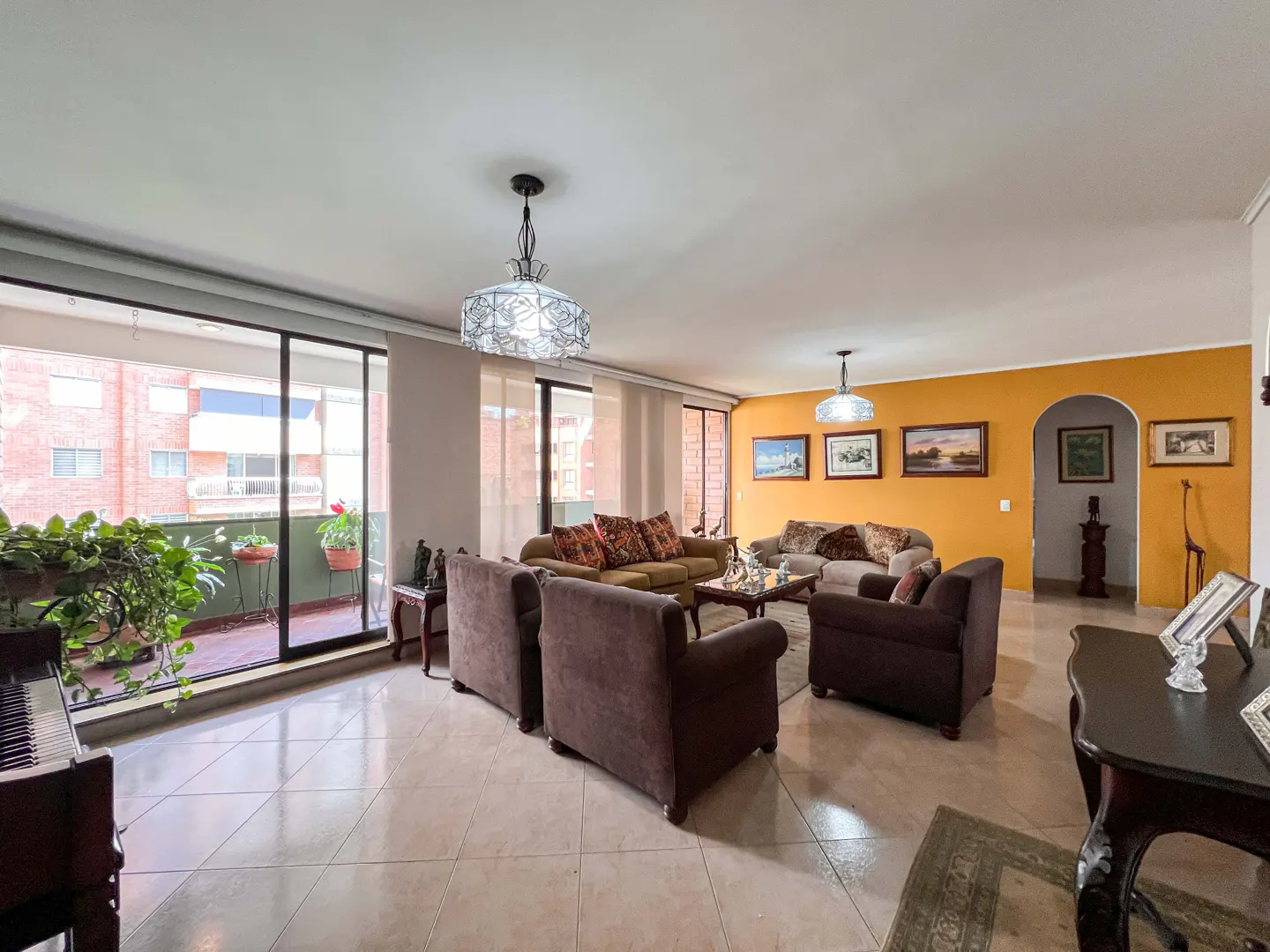 A spacious living room with brown sofas, armchairs, and a piano by a balcony with plants. The walls are yellow, adorned with framed artwork.