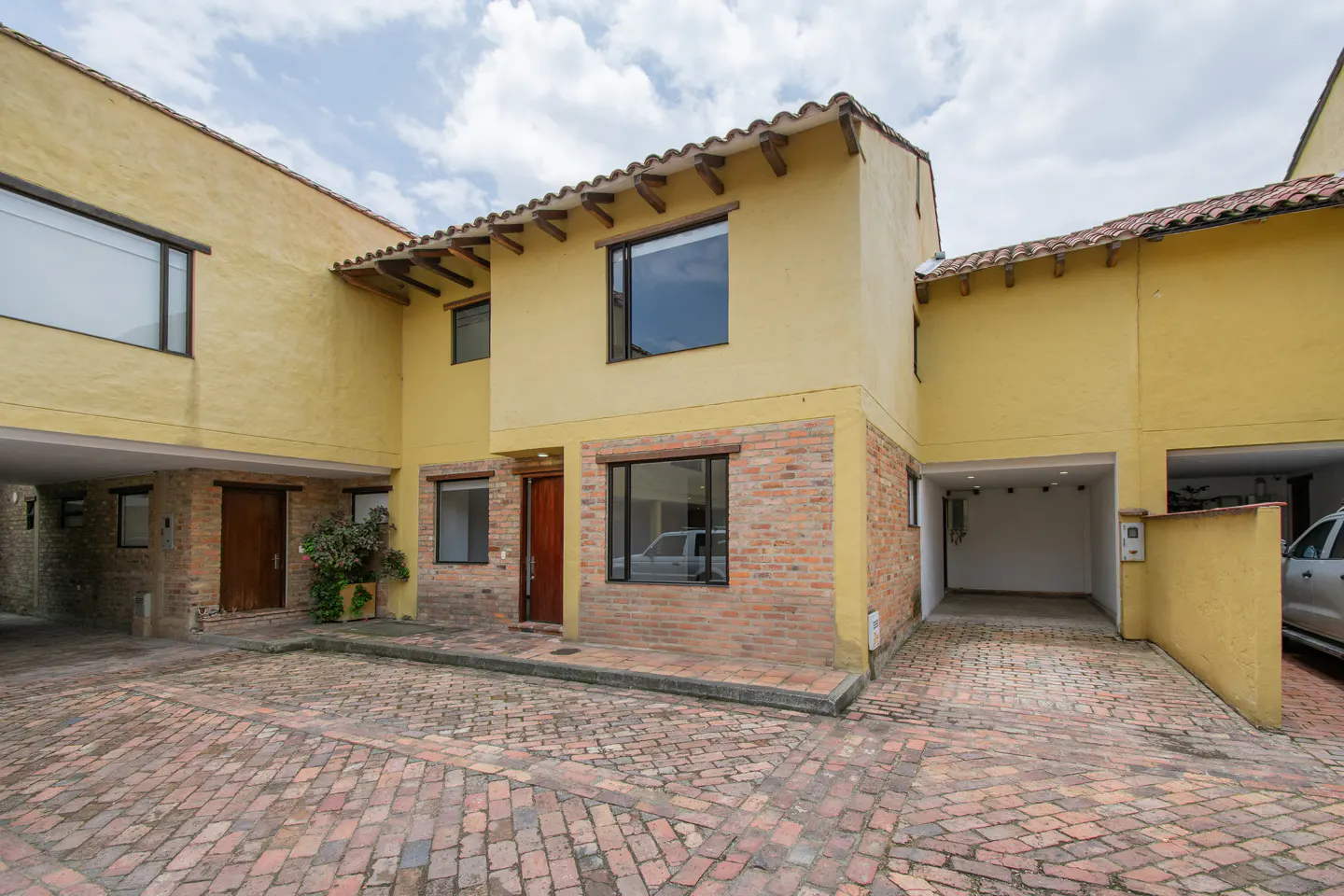 Exterior of a yellow two-story townhouse with brick accents and a brick driveway. A car is visible in the garage.