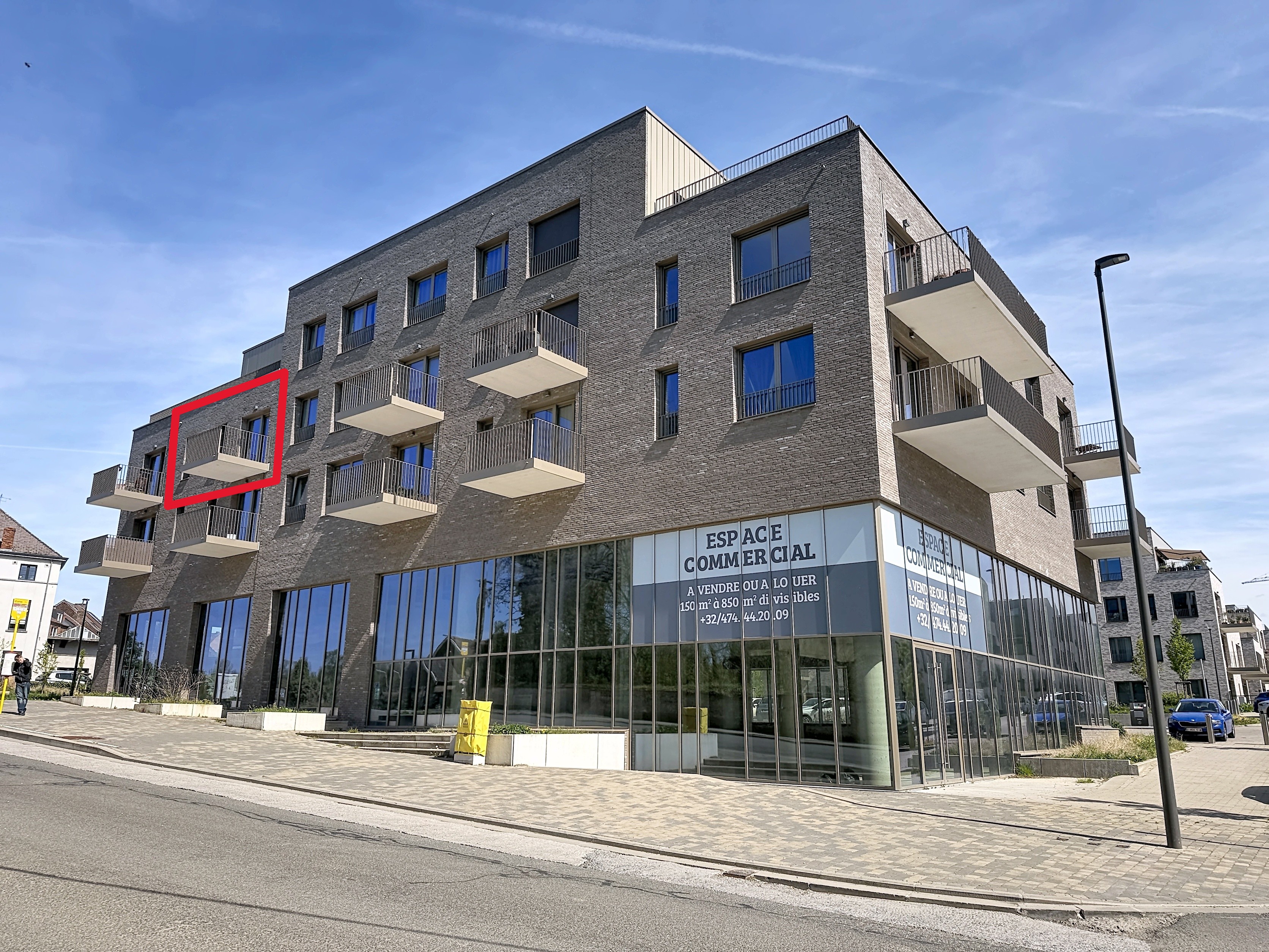 Modern gray brick building with balconies and commercial space on the ground floor. Red box highlights a balcony.