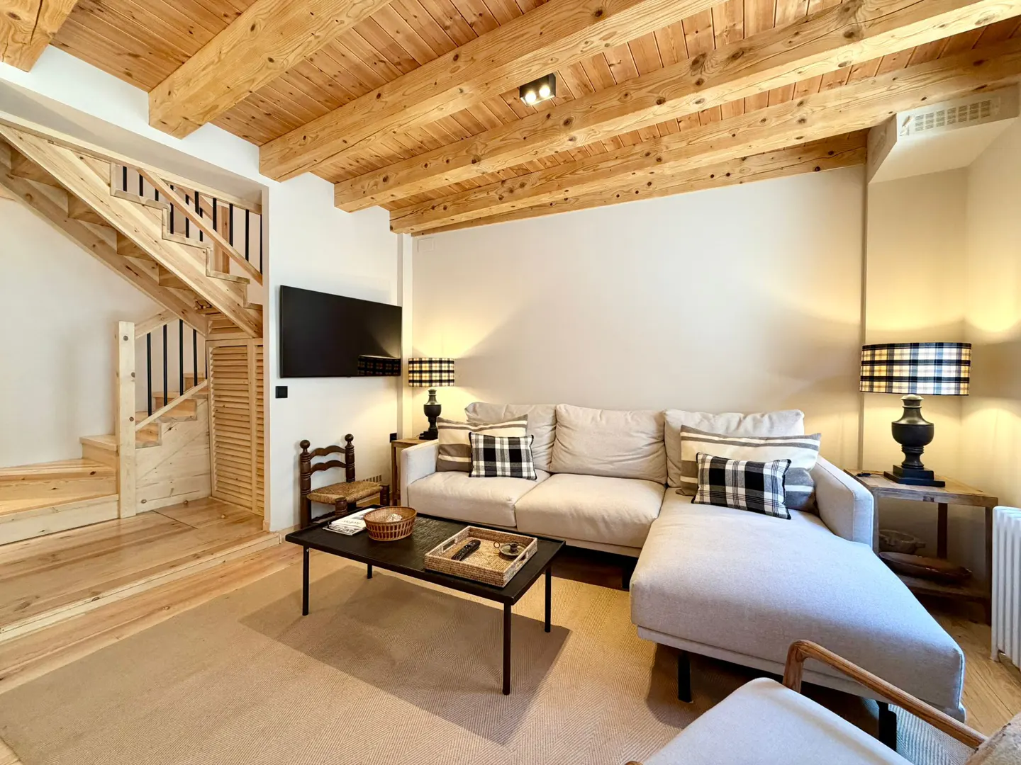 Living room with wooden ceiling beams, a beige sectional sofa with plaid pillows, and a black coffee table. A wooden staircase is on the left.