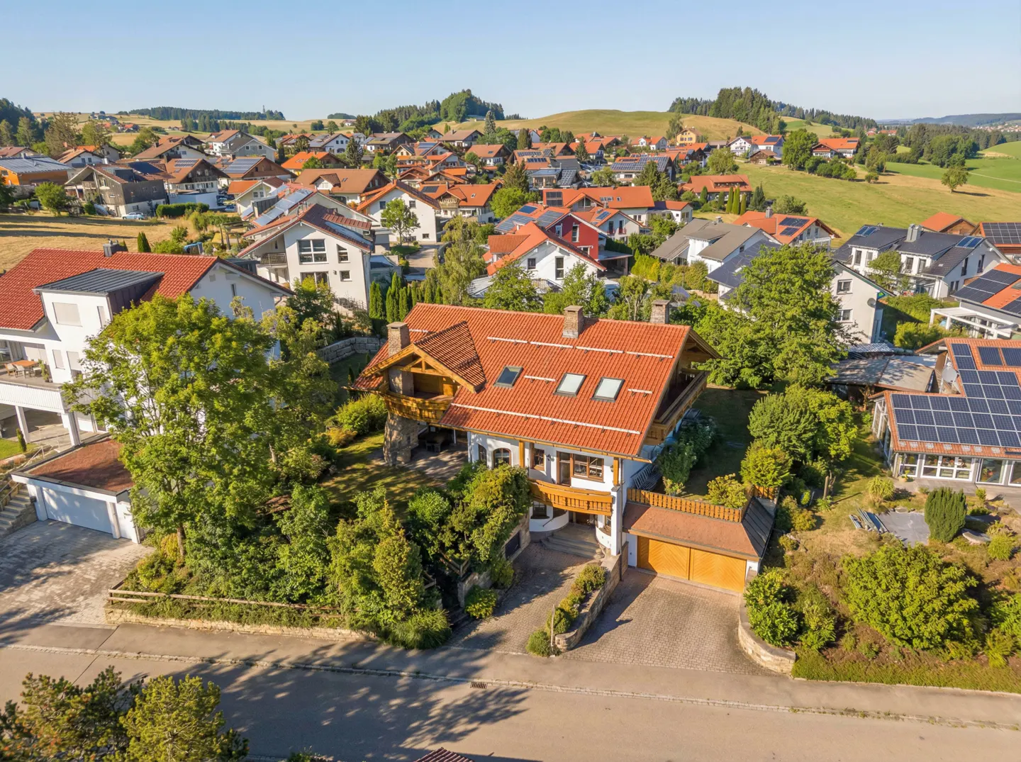 Aerial view of a white house with a red tile roof, surrounded by trees and other houses in a rural setting.