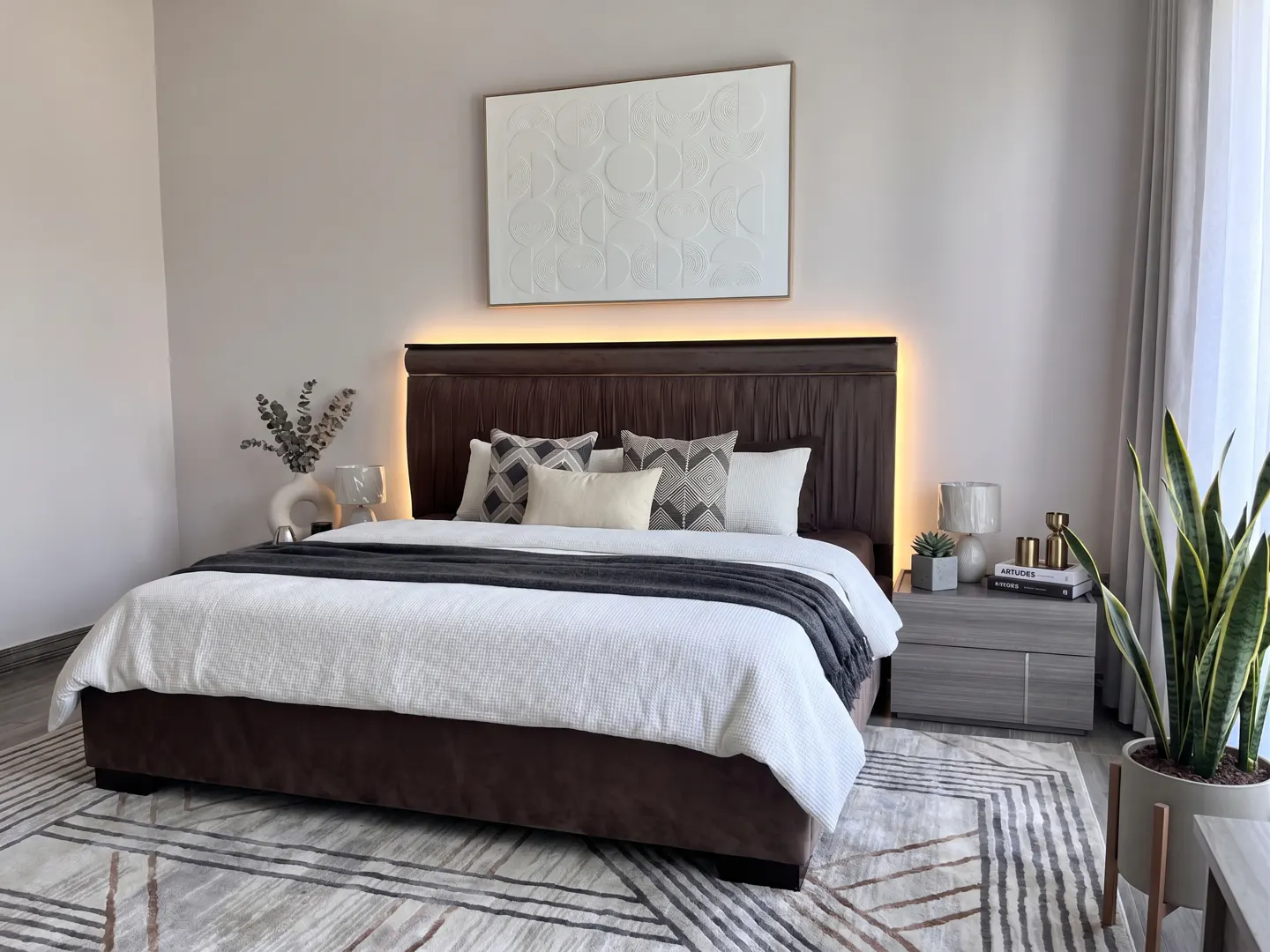Bedroom with a brown bed, white bedding, and decorative pillows. A white textured art piece hangs above the headboard with warm lighting. A gray nightstand and potted plant are visible.