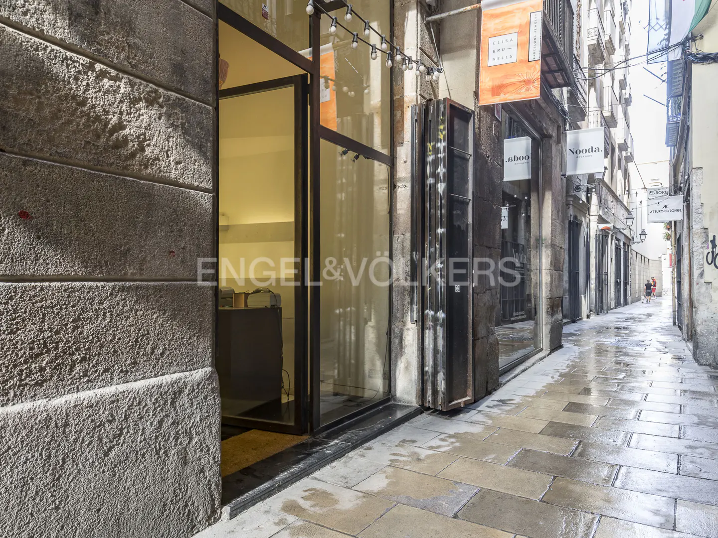 Exterior view of a narrow street with shops, featuring stone buildings and a wet, tiled walkway.
