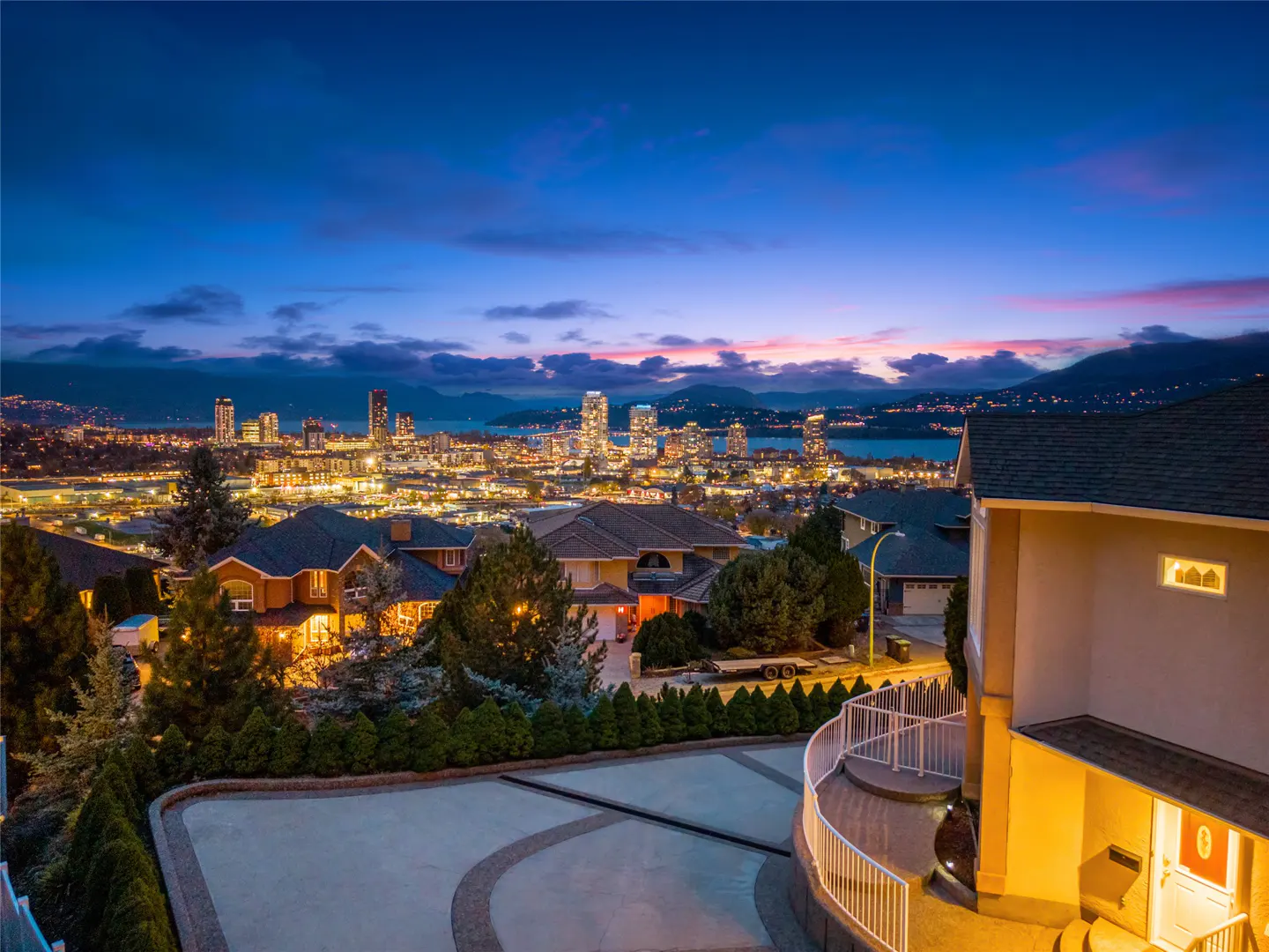 Night view of a house with a circular driveway, overlooking a city skyline with illuminated buildings and a lake.
