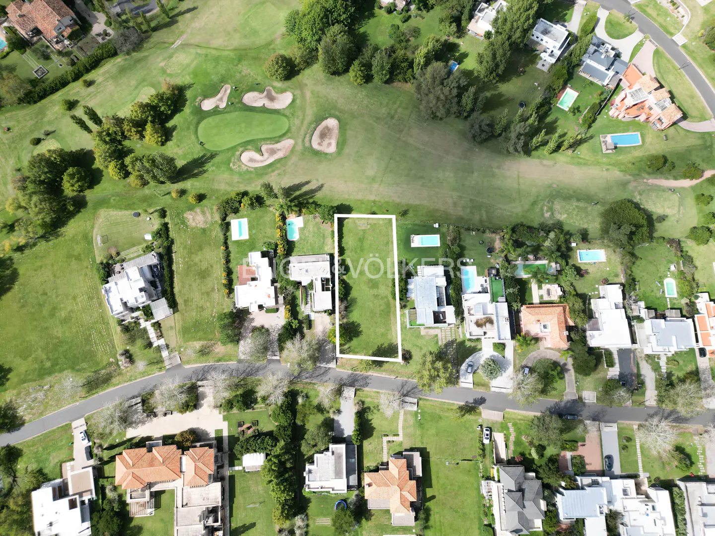 Aerial view of a rectangular grassy lot outlined in white, surrounded by houses, trees, and a golf course.