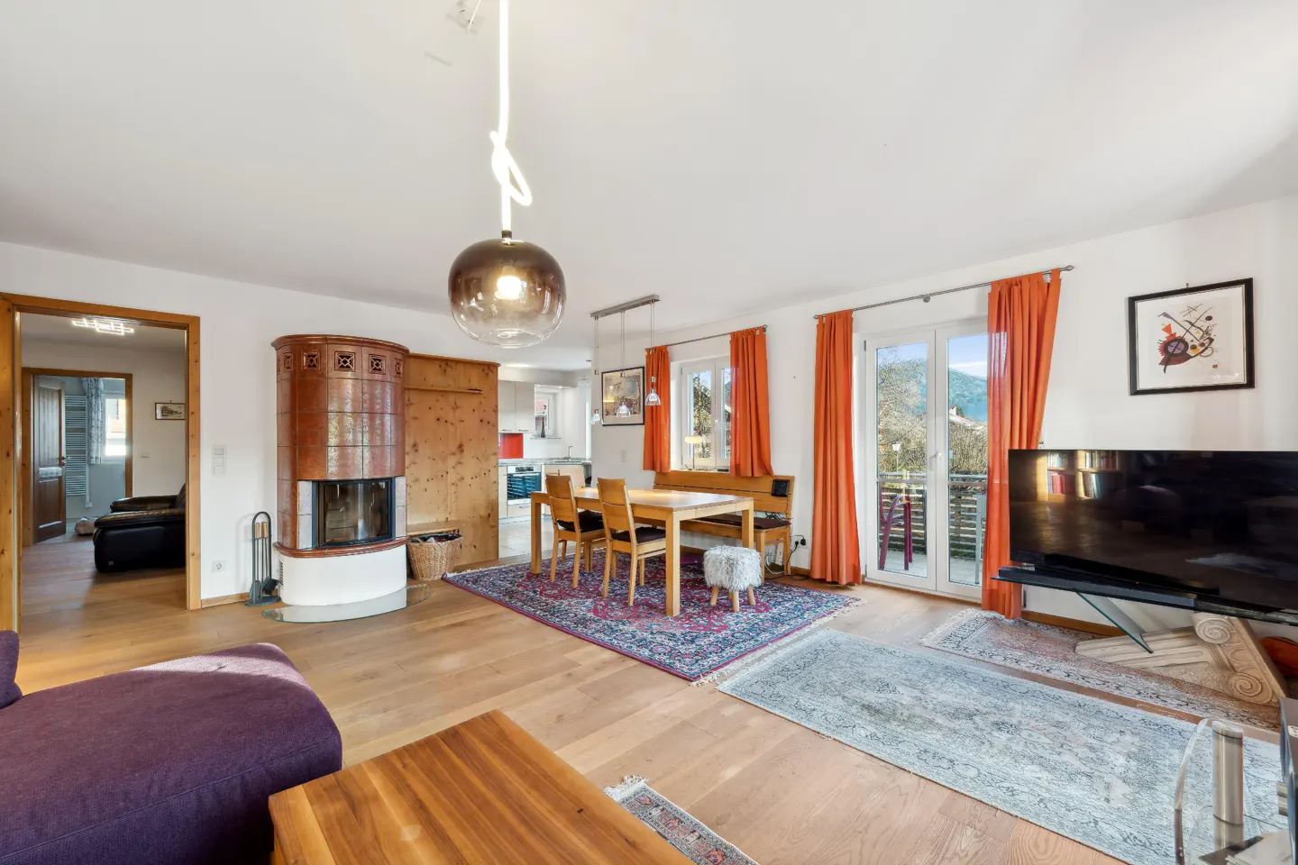 Bright living room with wood floors, a dining table, and a fireplace. Orange curtains frame the balcony doors. A purple sofa is in the foreground.