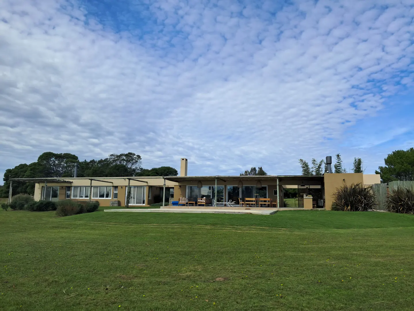 A tan house with a chimney sits on a green lawn under a cloudy sky. Trees are visible in the background.