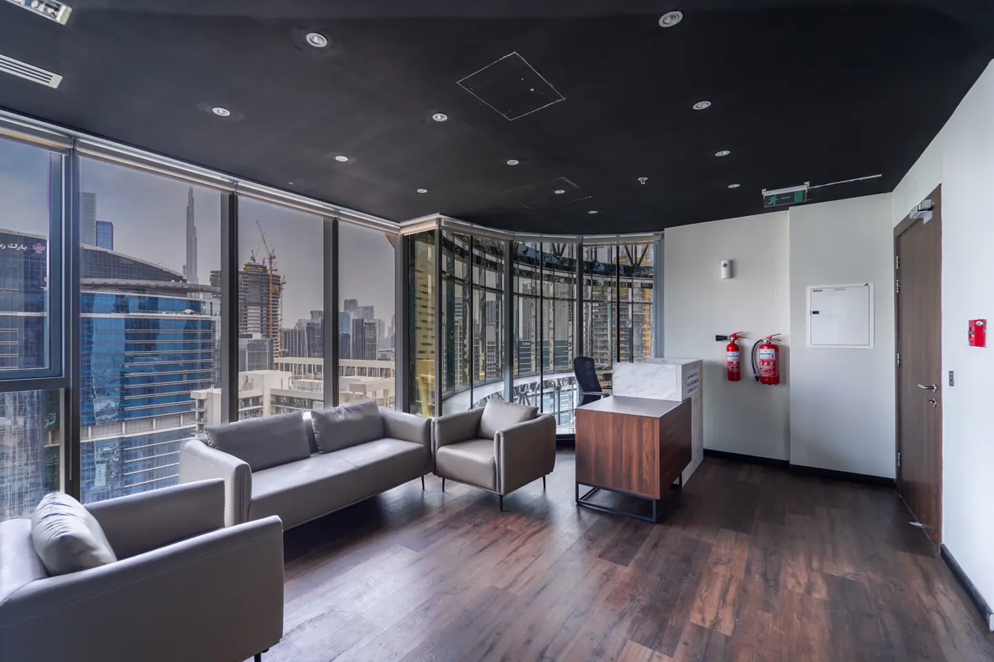 Modern office lobby with gray sofas, wood floors, and a reception desk. Large windows offer a city view. Fire extinguishers are mounted on the wall.