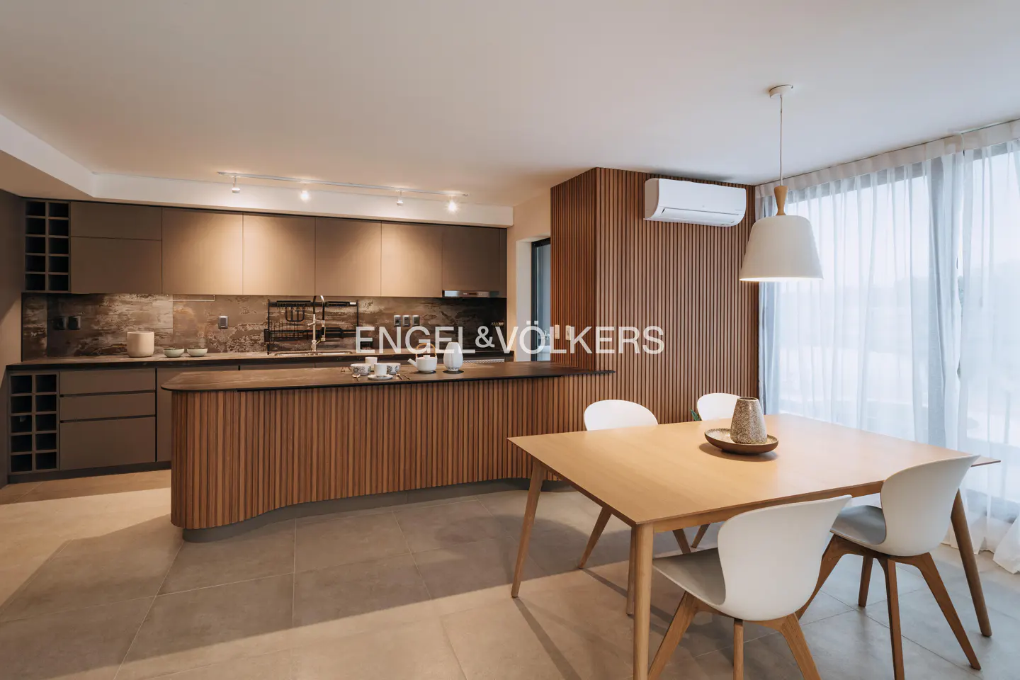 Open-concept kitchen and dining area with wood accents, gray cabinets, and a light wood table with white chairs. Natural light filters through sheer curtains.