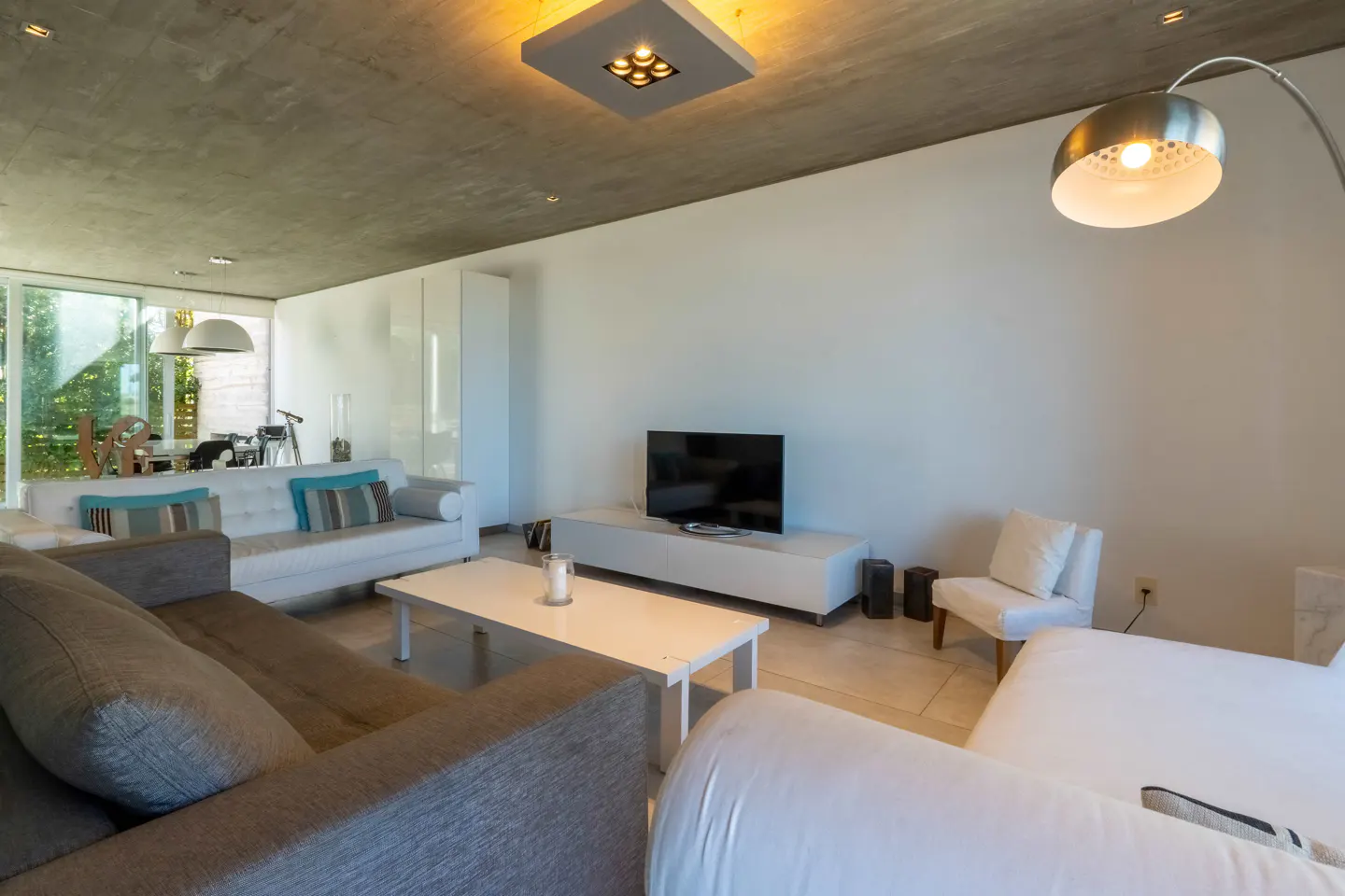 Modern living room with gray sofa, white couches, TV, and a silver floor lamp. The ceiling is concrete with a square light fixture.