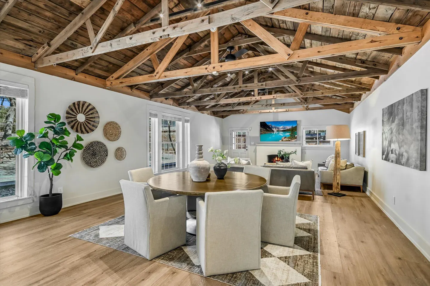 Open-concept living and dining area with exposed wood beams, white walls, and light wood floors. Round dining table with six chairs. Fireplace and TV in the background.
