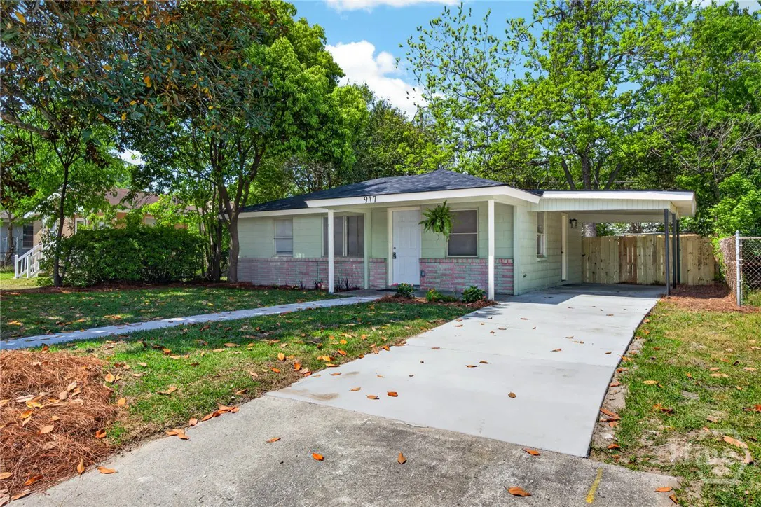 A light green single-story house with a carport, white trim, and a gray driveway. Trees surround the house.