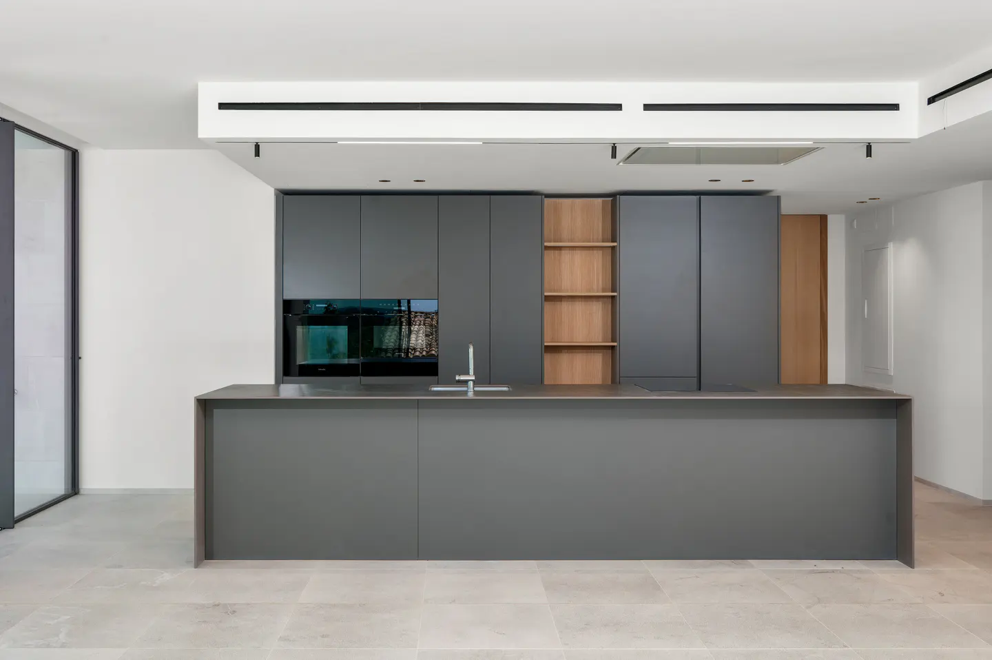 Modern kitchen with gray cabinets, wood shelving, and a large island. Stainless steel sink and built-in oven. Light tile flooring.
